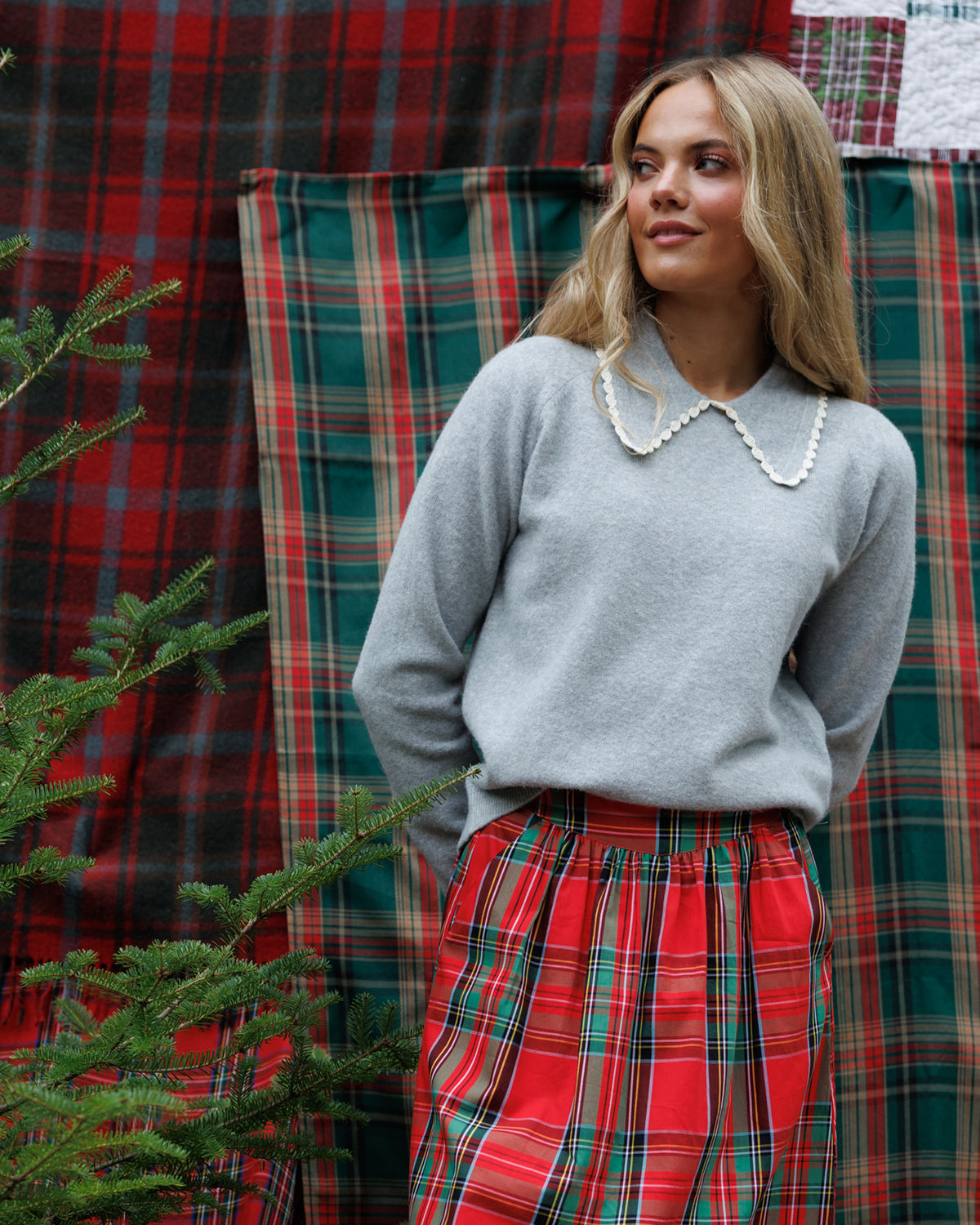 A woman wearing a light gray knit sweater with a pearl collar and a red tartan skirt, standing in front of a red and green plaid fabric backdrop with a small Christmas tree in the foreground.