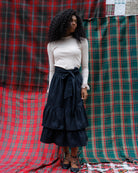 A woman stands in front of a red and green plaid textile backdrop, wearing a cream ribbed long-sleeve top, a dark navy tiered skirt with a side bow, and black lace-up flats.