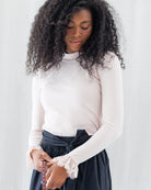 Front view of a light blush pink ribbed long-sleeve mock-neck top featuring delicate scalloped eyelash lace trim at the flared cuffs.