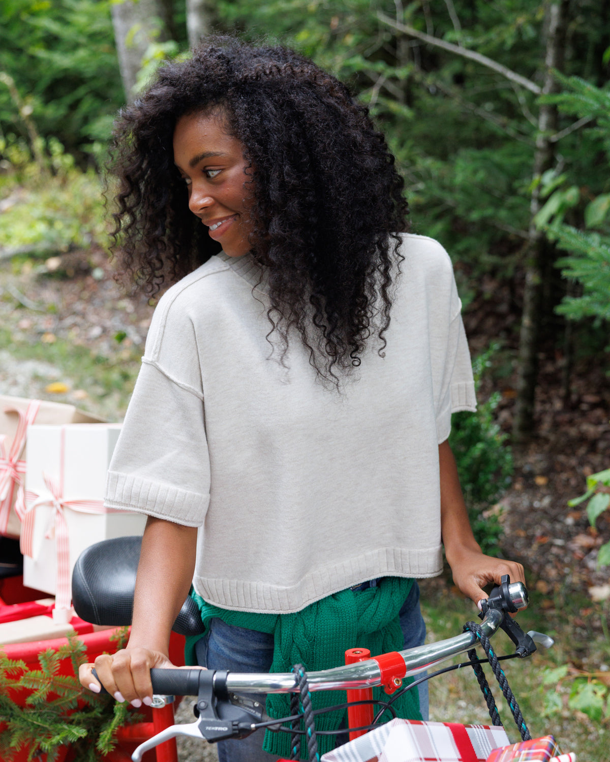 A woman with long, curly black hair stands outdoors next to a bicycle, wearing a light beige knit short-sleeve top and a green sweater tied around her waist. Gift boxes are visible in the background.