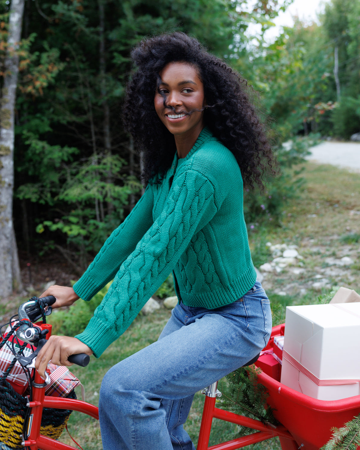 A woman with curly black hair smiles while riding a red cargo bike outdoors on a wooded path. She is wearing a green cable-knit sweater and blue jeans, with a white gift box in the bike's red rear basket.