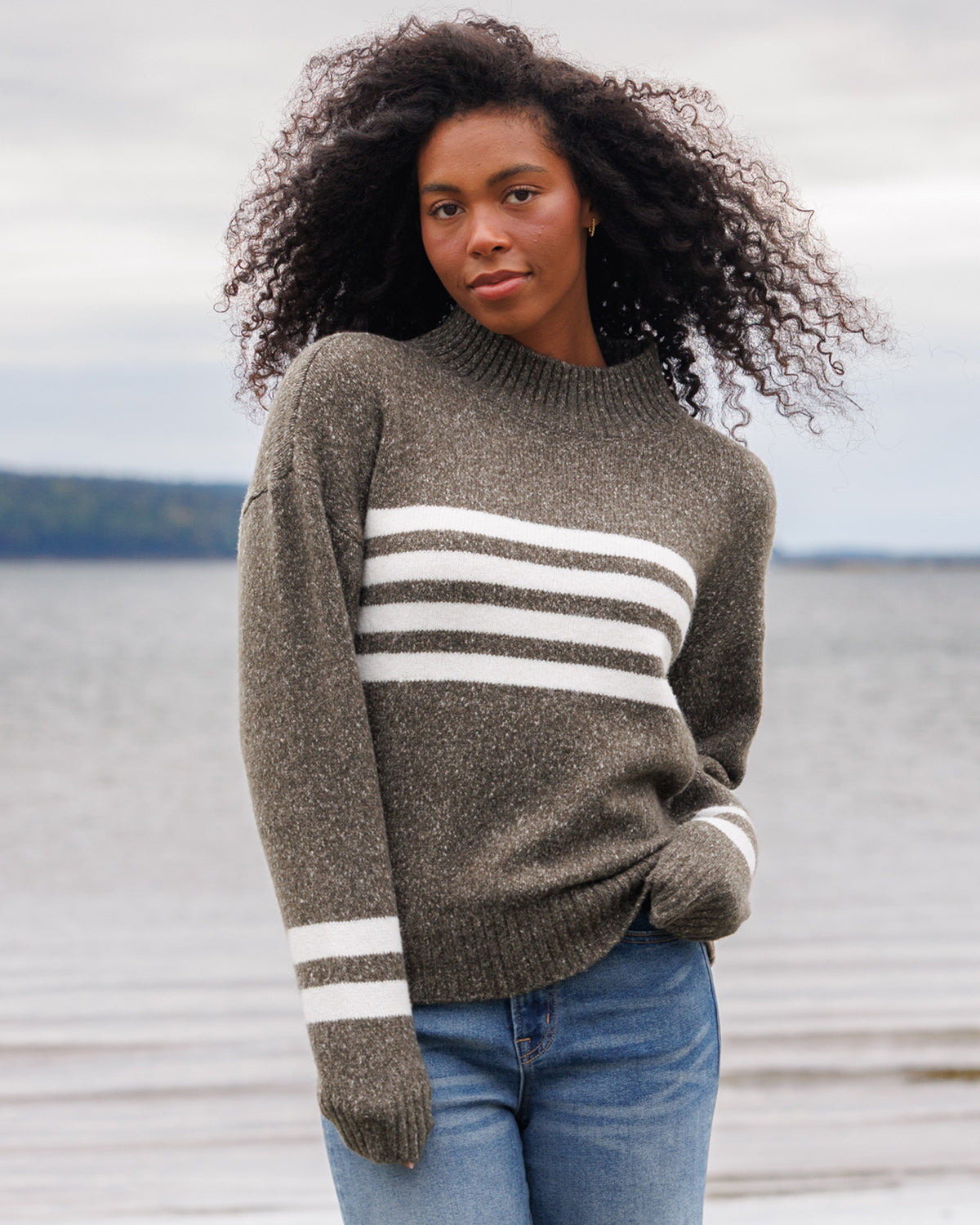 A woman with curly dark hair stands outdoors by the water wearing a heathered taupe knit sweater with three horizontal white stripes across the chest and white stripes on the sleeves, paired with blue jeans.