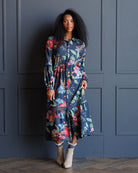 A woman with curly black hair stands against a dark blue paneled wall wearing a long-sleeved navy blue midi dress with a colorful floral print, paired with beige ankle boots.