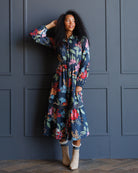 A woman with curly black hair models a long-sleeved midi dress with a colorful floral print on a dark navy background. She is standing in front of a dark gray paneled wall, wearing beige ankle boots.