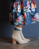 Beige suede ankle boots with a stacked wooden block heel, worn with a navy floral skirt and a white lace sock, standing on a dark wood floor against a dark blue-gray wall.