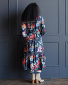 A woman viewed from behind wearing a navy blue midi dress with a vibrant multicolor floral print, paired with beige ankle boots, standing in front of a dark navy paneled wall.