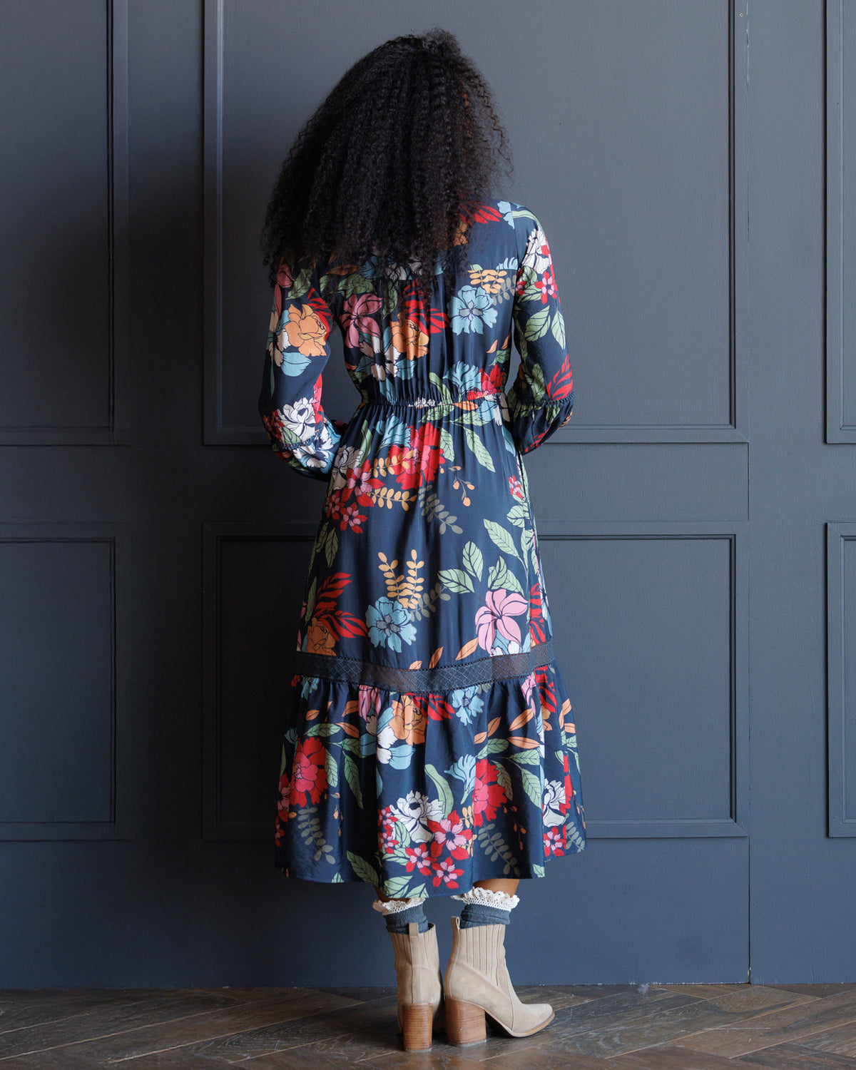 A woman viewed from behind wearing a navy blue midi dress with a vibrant multicolor floral print, paired with beige ankle boots, standing in front of a dark navy paneled wall.