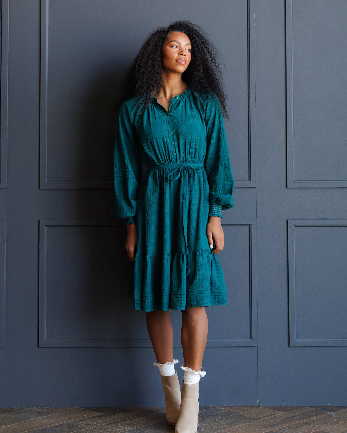 A woman with curly black hair stands against a dark blue paneled wall wearing a teal green long-sleeve midi dress with a tie waist and cream ankle boots.