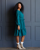 A woman wearing a teal green tiered midi dress with long puff sleeves and a tie waist, paired with beige ankle boots and white ruffled socks, posing against a dark blue paneled wall.