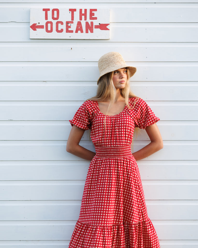 Woman in a red checkered dress standing in front of a wooden wall with 'To The Ocean' sign.