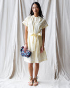 Woman wearing a light yellow dress with a belt, holding a clutch, against a white curtain background