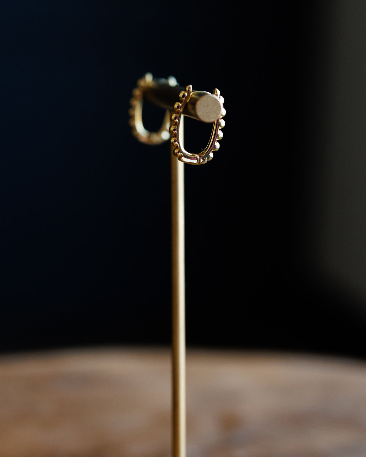 A pair of gold hoop earrings with small bead details along the front edge, displayed on a gold stand against a dark, blurred background.