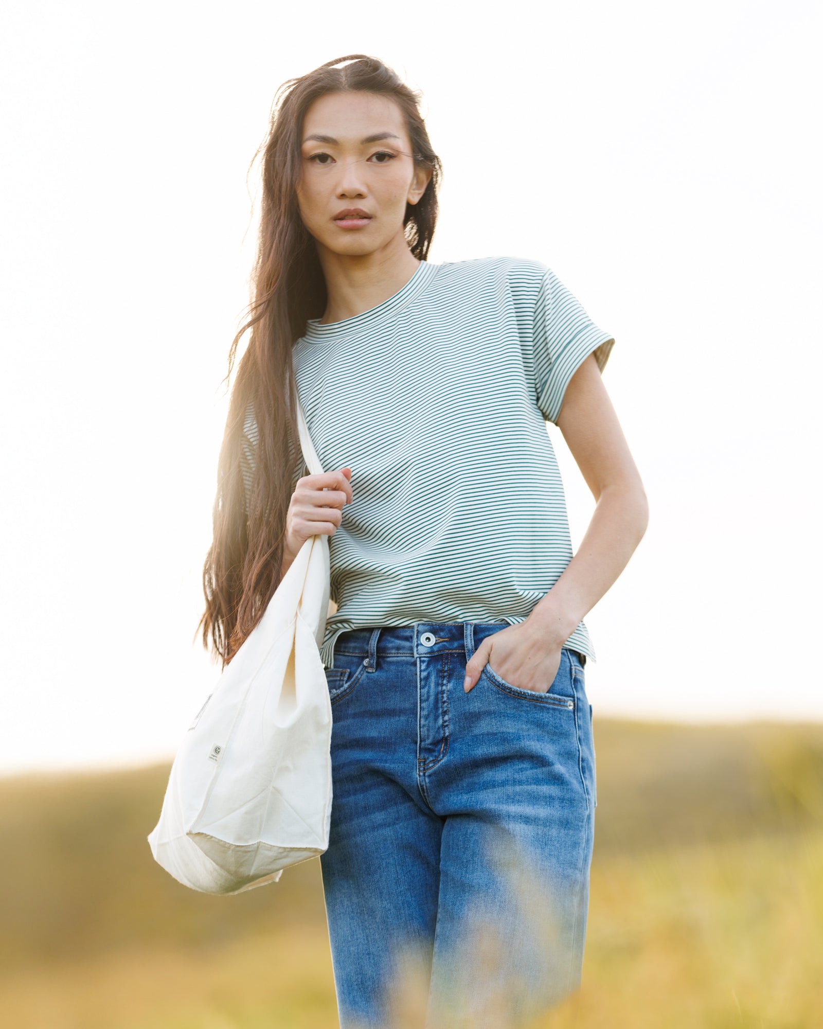 Woman wearing a striped t-shirt and jeans in a field and holding a bag