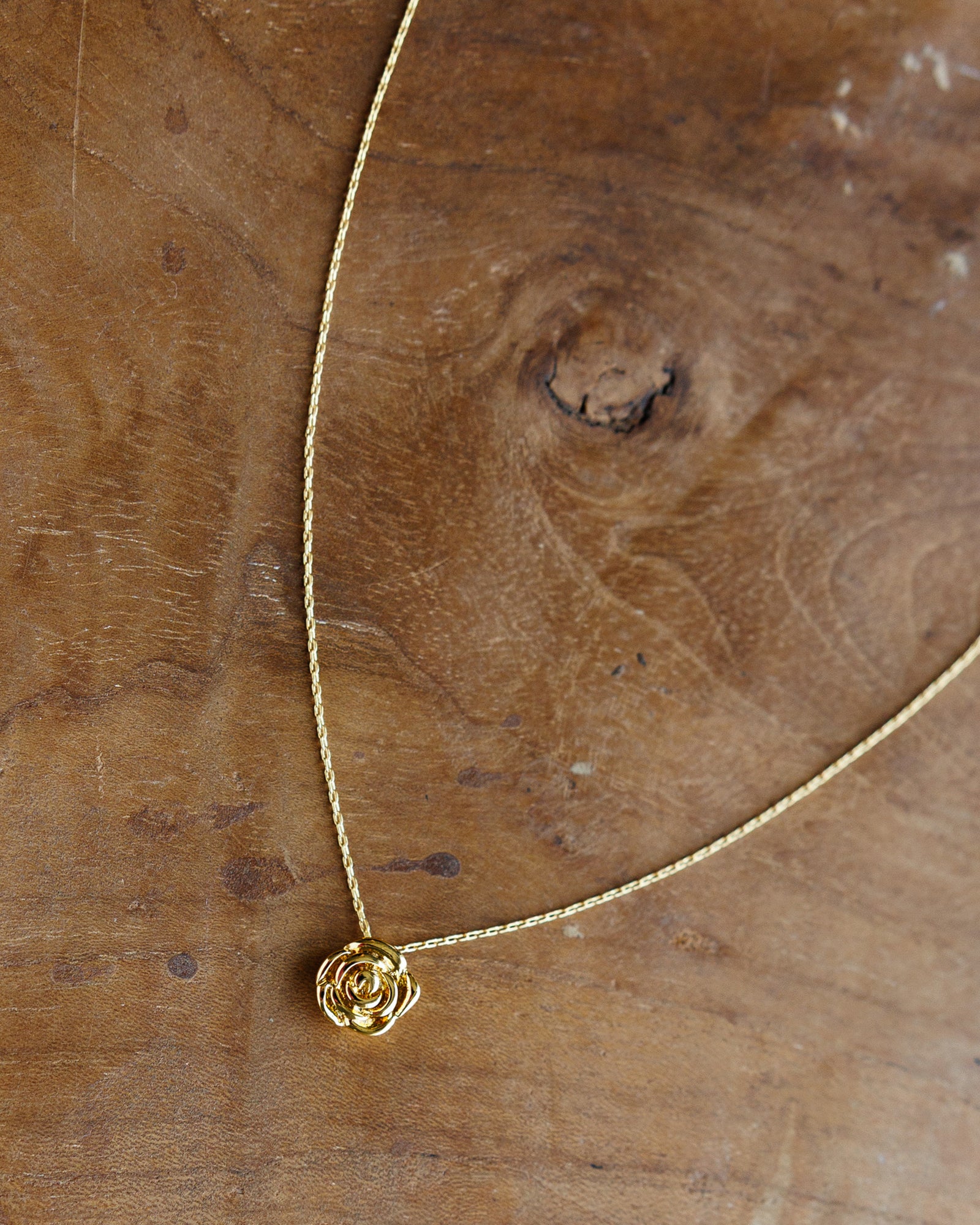 A delicate gold chain necklace with a small rose-shaped pendant, displayed on a rustic wooden surface.