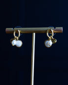 Gold-tone dangle earrings with two white pearl-like beads and small gold charms, displayed on a gold stand against a dark navy background.