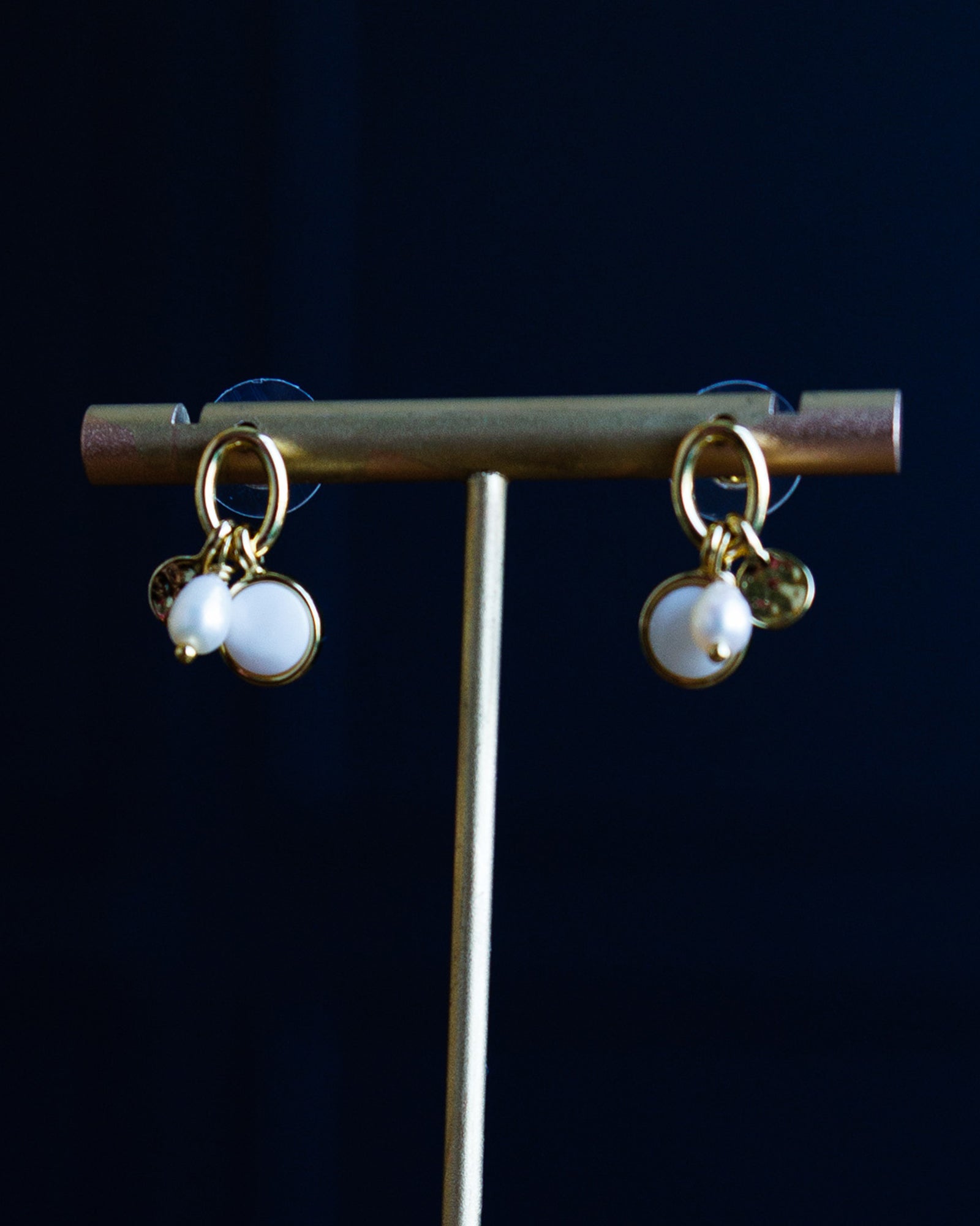 Gold-tone dangle earrings with two white pearl-like beads and small gold charms, displayed on a gold stand against a dark navy background.