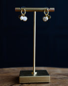 A pair of gold dangle earrings with white pearl-like beads, displayed on a gold stand against a dark navy background.