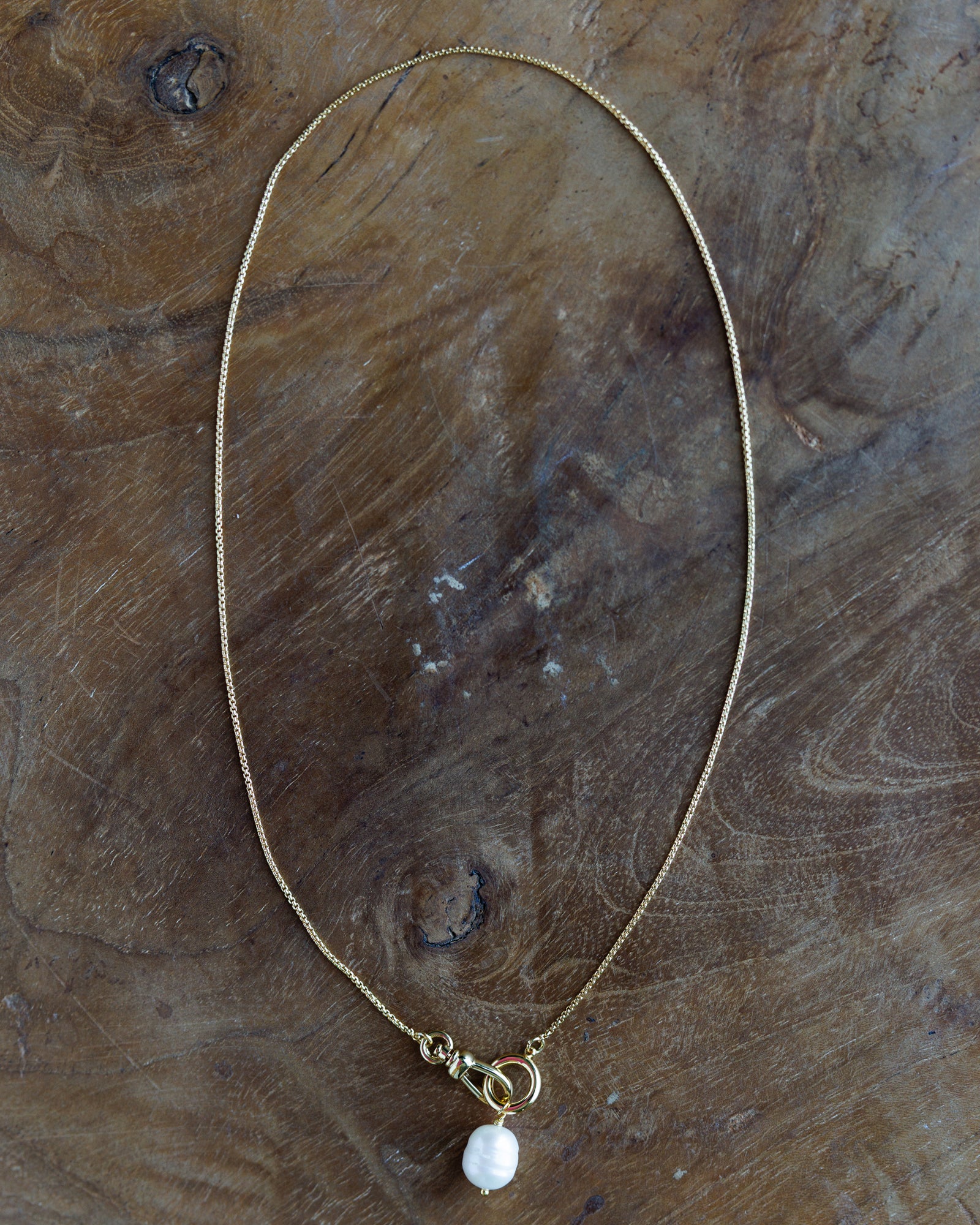 A delicate gold chain necklace with a small white baroque pearl pendant, displayed on a rustic, textured wooden surface.