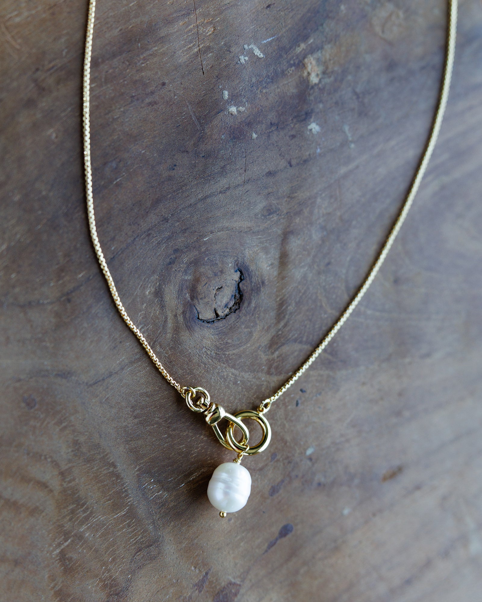 A delicate gold chain necklace with a unique clasp featuring a small white baroque pearl, displayed on a rustic wooden surface.