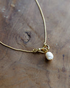 A delicate gold chain necklace featuring a lobster clasp and a single small white baroque pearl pendant, displayed on a rustic wooden surface.