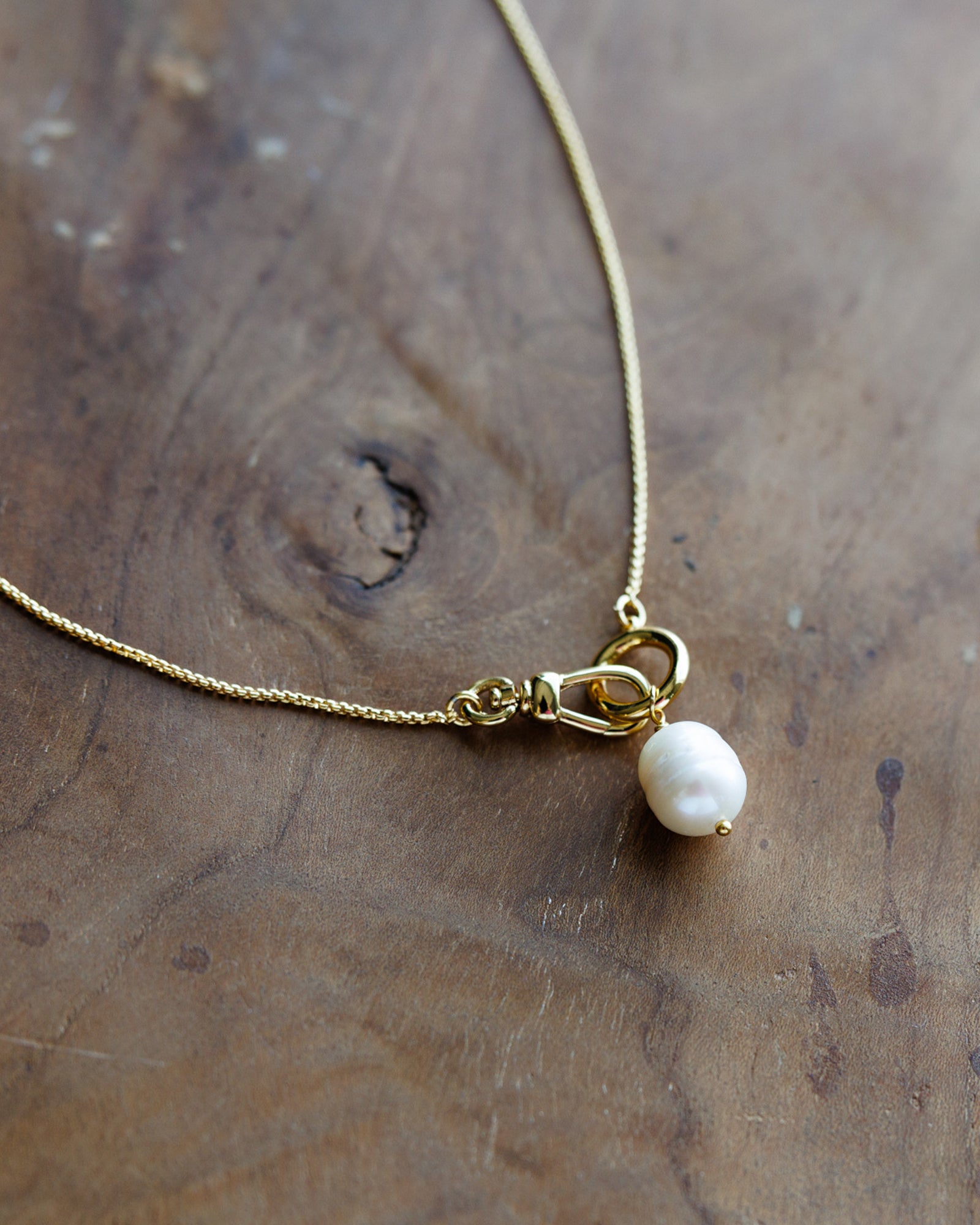 A delicate gold chain necklace featuring a lobster clasp and a single small white baroque pearl pendant, displayed on a rustic wooden surface.