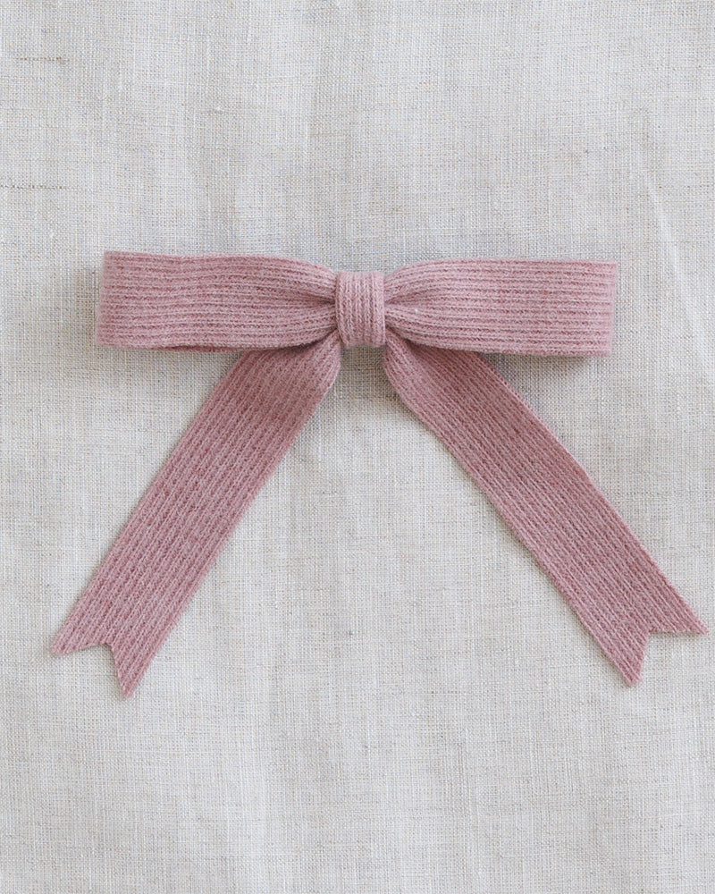 A dusty pink ribbed knit bow accessory, styled on a textured beige linen fabric background.