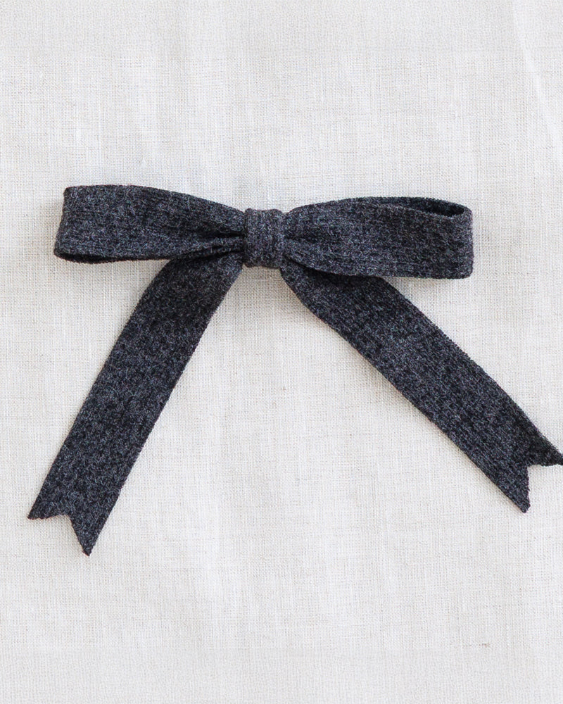 A dark charcoal gray textured fabric bow, neatly tied and displayed flat against a light beige linen background.