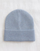 A light blue knit beanie hat with a folded cuff, laid flat on a textured off-white fabric background.