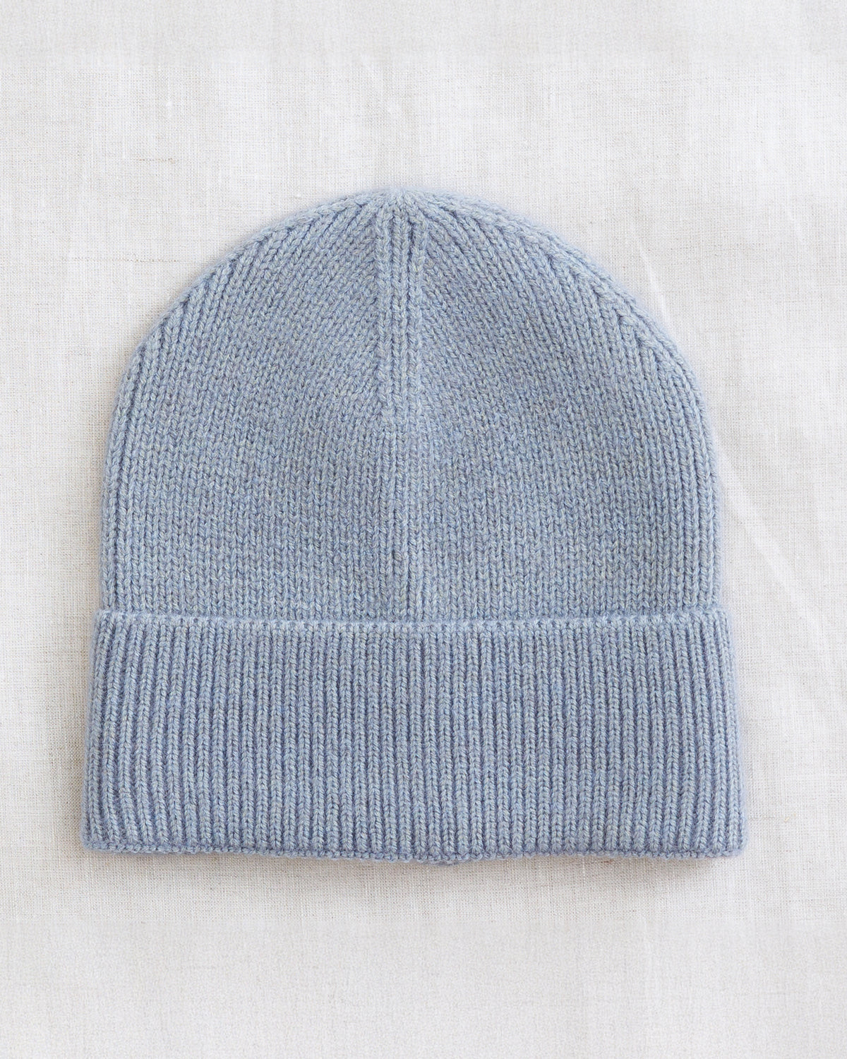 A light blue knit beanie hat with a folded cuff, laid flat on a textured off-white fabric background.