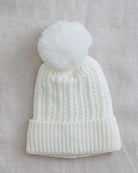 An off-white cable-knit beanie with a matching fluffy pom-pom, laid flat on a textured beige linen background.