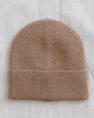 A light brown ribbed knit beanie hat laid flat on a textured off-white linen background.