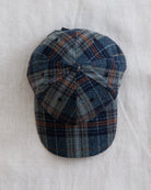 A classic six-panel flat-brim cap in a blue, gray, and rust-orange plaid pattern, laid flat on a textured off-white linen background.