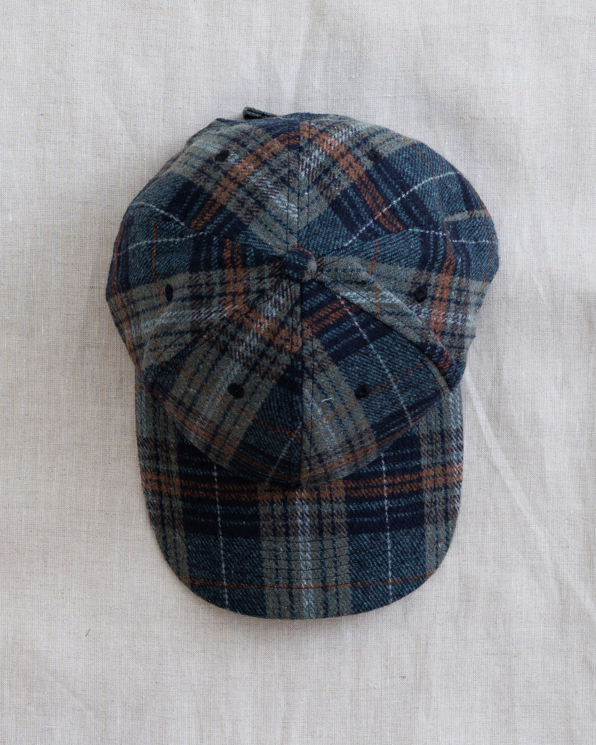 A classic six-panel flat-brim cap in a blue, gray, and rust-orange plaid pattern, laid flat on a textured off-white linen background.