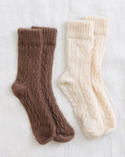 Three pairs of cable-knit socks laid flat on a light fabric background: one medium brown, one cream, and one warm off-white.
