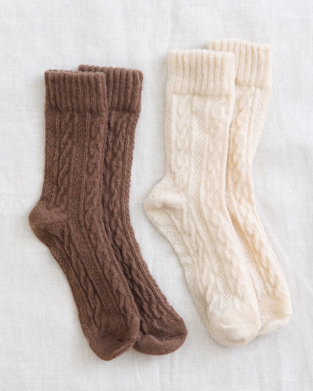 Three pairs of cable-knit socks laid flat on a light fabric background: one medium brown, one cream, and one warm off-white.