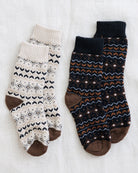 Two pairs of patterned crew socks laid flat on a white fabric background. The left pair is cream with black geometric patterns and brown heels, toes, and cuffs.