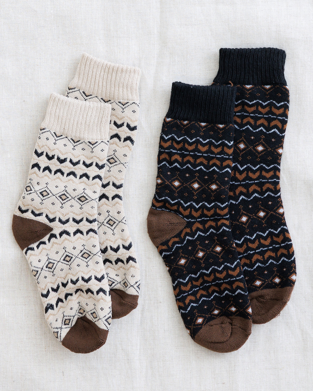 Two pairs of patterned crew socks laid flat on a white fabric background. The left pair is cream with black geometric patterns and brown heels, toes, and cuffs.