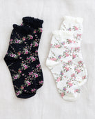 Two pairs of floral ruffled socks laid flat on a light fabric background. One pair is black with pink and green floral print, and the other pair is white with a matching pink and green floral print.