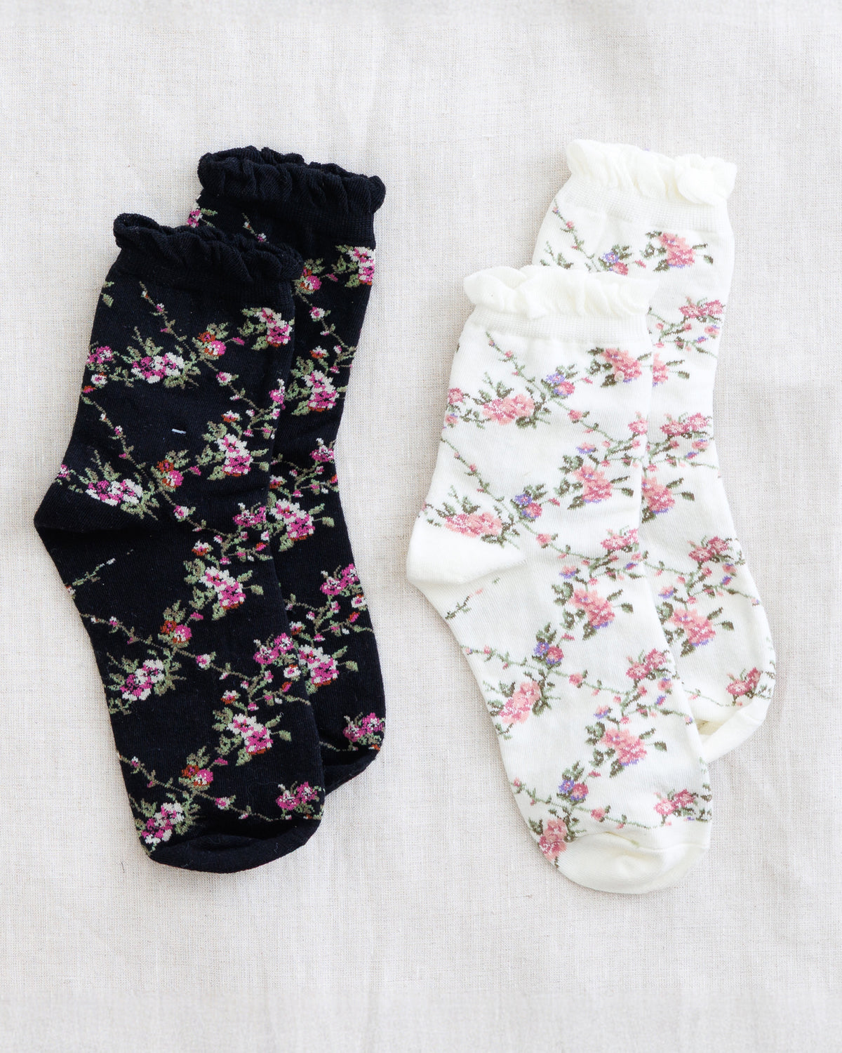 Two pairs of floral ruffled socks laid flat on a light fabric background. One pair is black with pink and green floral print, and the other pair is white with a matching pink and green floral print.