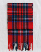 A folded red tartan plaid scarf with fringed ends, featuring navy, white, and thin pink stripes, laid flat on a white textured fabric background.