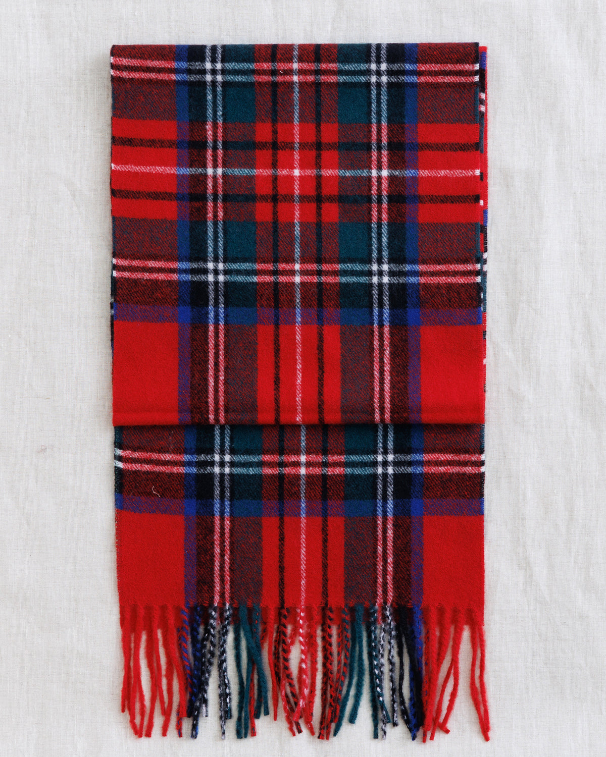 A folded red tartan plaid scarf with fringed ends, featuring navy, white, and thin pink stripes, laid flat on a white textured fabric background.
