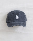 A dark gray washed cotton baseball cap featuring a small white embroidered ghost design on the front, displayed flat against a light beige textured fabric background.
