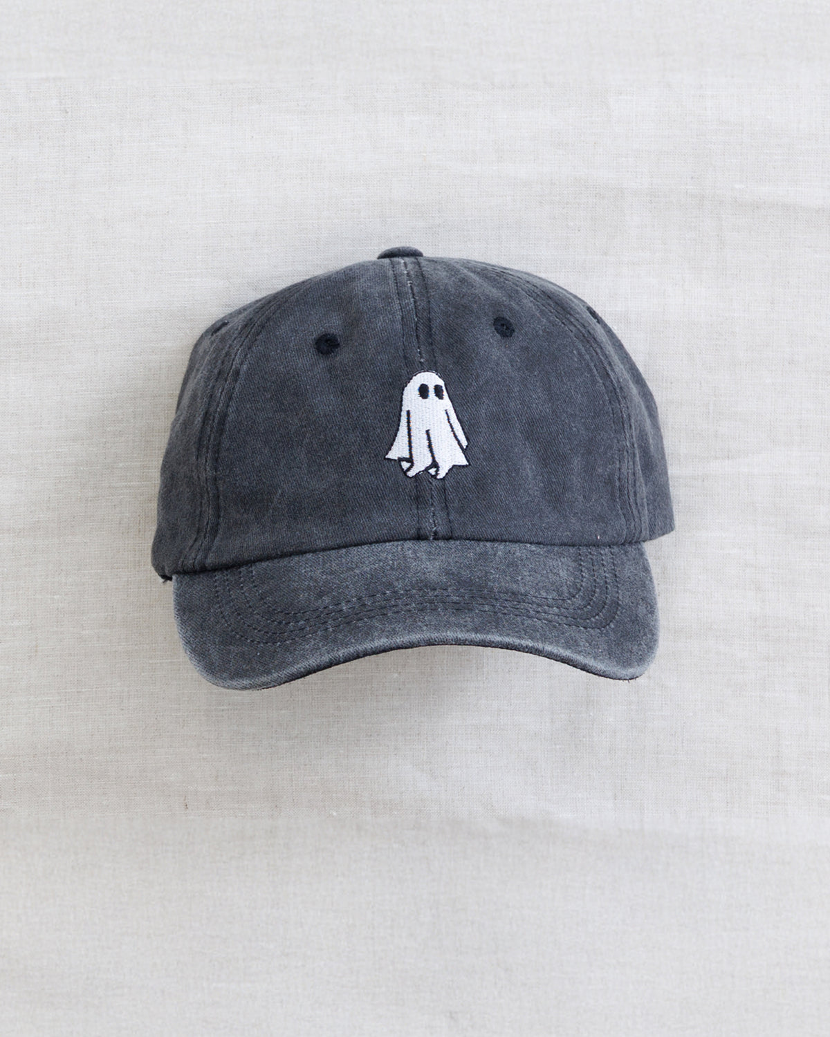 A dark gray washed cotton baseball cap featuring a small white embroidered ghost design on the front, displayed flat against a light beige textured fabric background.