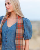 A woman wearing a blue denim dress with puff sleeves and a red, orange, and beige plaid scarf draped around her neck, photographed outdoors against a soft-focus sandy background.