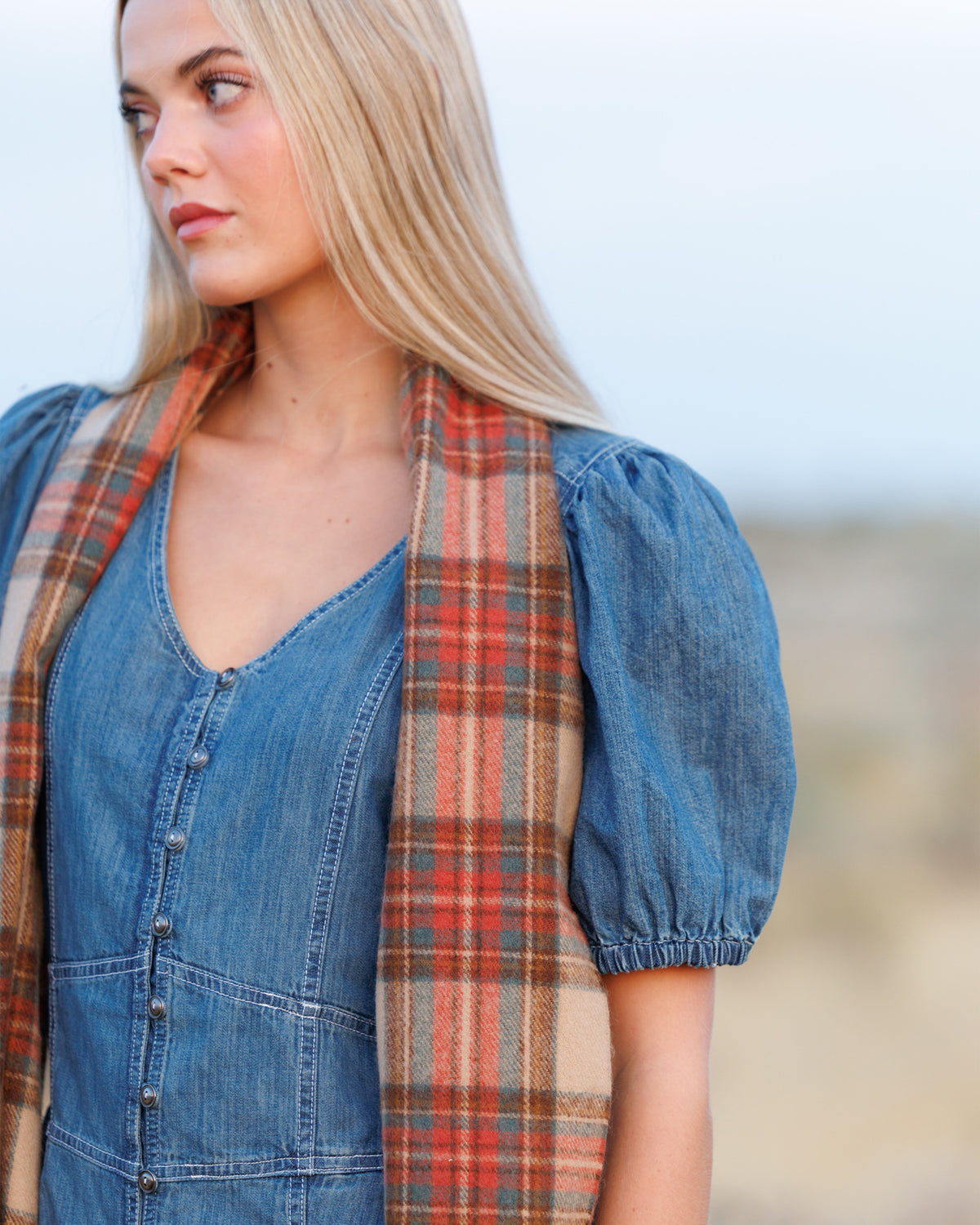 A woman wearing a blue denim dress with puff sleeves and a red, orange, and beige plaid scarf draped around her neck, photographed outdoors against a soft-focus sandy background.
