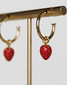 Gold hoop earrings with red strawberry-shaped charms on a gold stand.