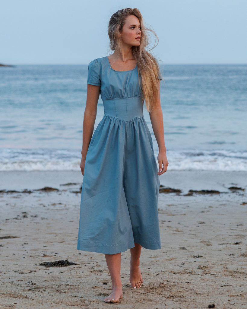 Woman in a blue dress standing on a beach with ocean in the background