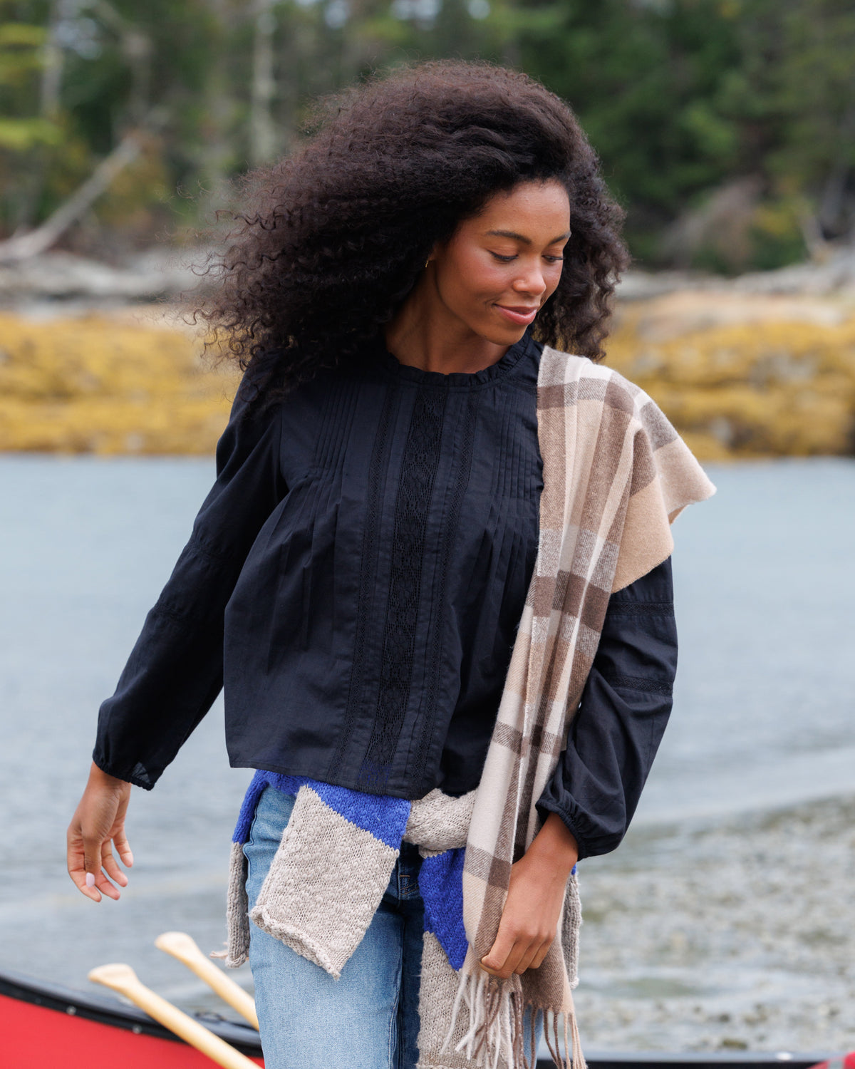 A woman with curly dark hair stands outdoors near water and trees, wearing a black long-sleeve blouse and a beige plaid shawl draped over her shoulders.