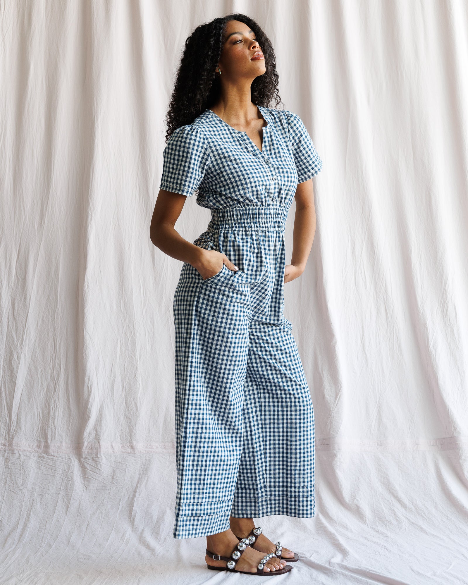 Woman wearing a blue and white checkered jumpsuit against a white curtain background