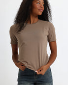 A woman wearing a brown short-sleeve crewneck t-shirt and jeans, standing against a fabric backdrop.